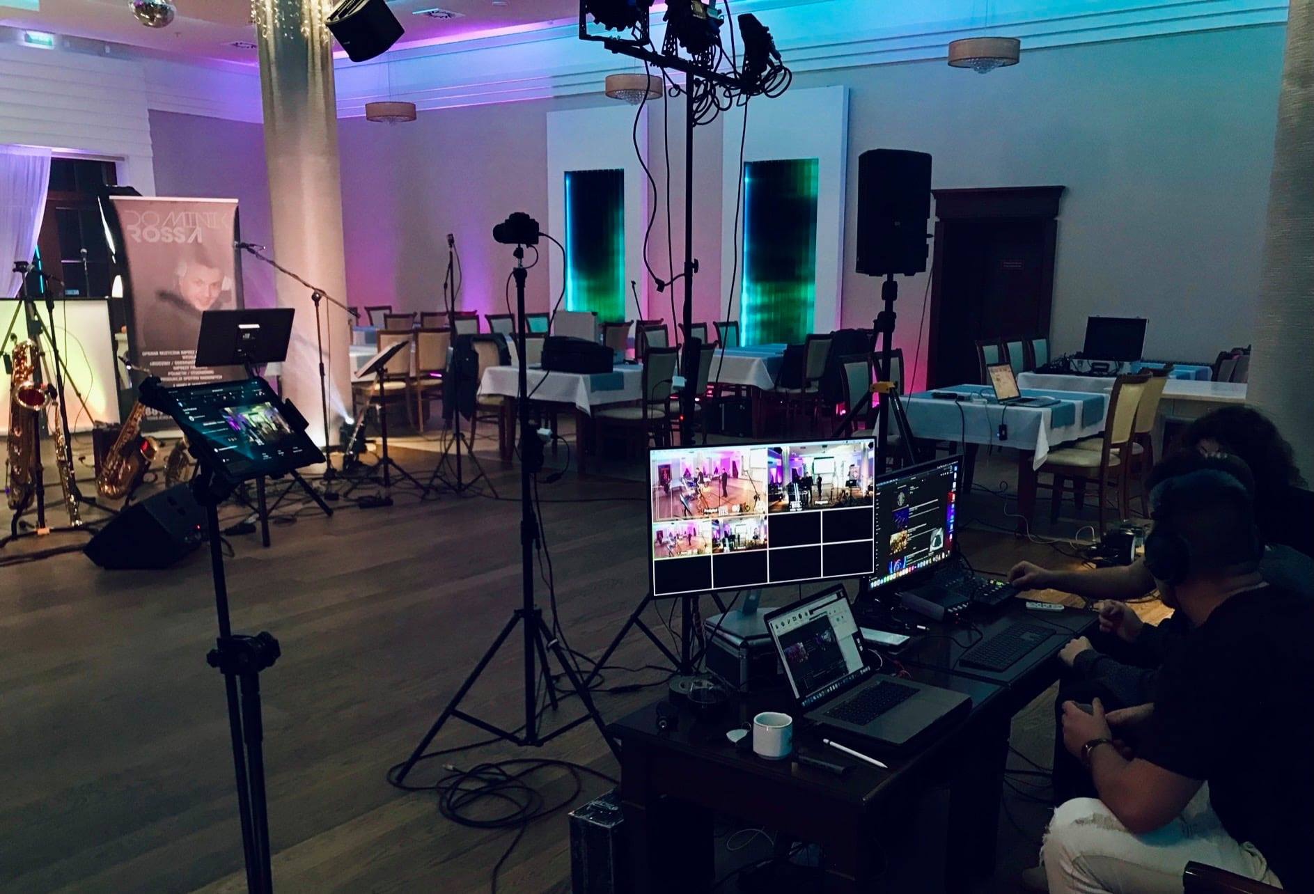 Sala bankietowa przygotowana do imprezy z rozstawionym sprzętem nagłośnieniowym, oświetleniowym i stanowiskiem do miksowania audio/wideo, widoczne monitory z podglądem kamer i laptop...