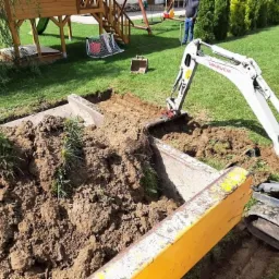 Mini koparka Takeuchi podczas prac ziemnych na trawniku, widoczny wykop z betonowym elementem, w tle plac zabaw i osoba.