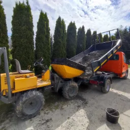 Pomarańczowa ciężarówka z żółtą przyczepą wyładowuje ziemię na placu budowy, w tle rząd wysokich tuj.