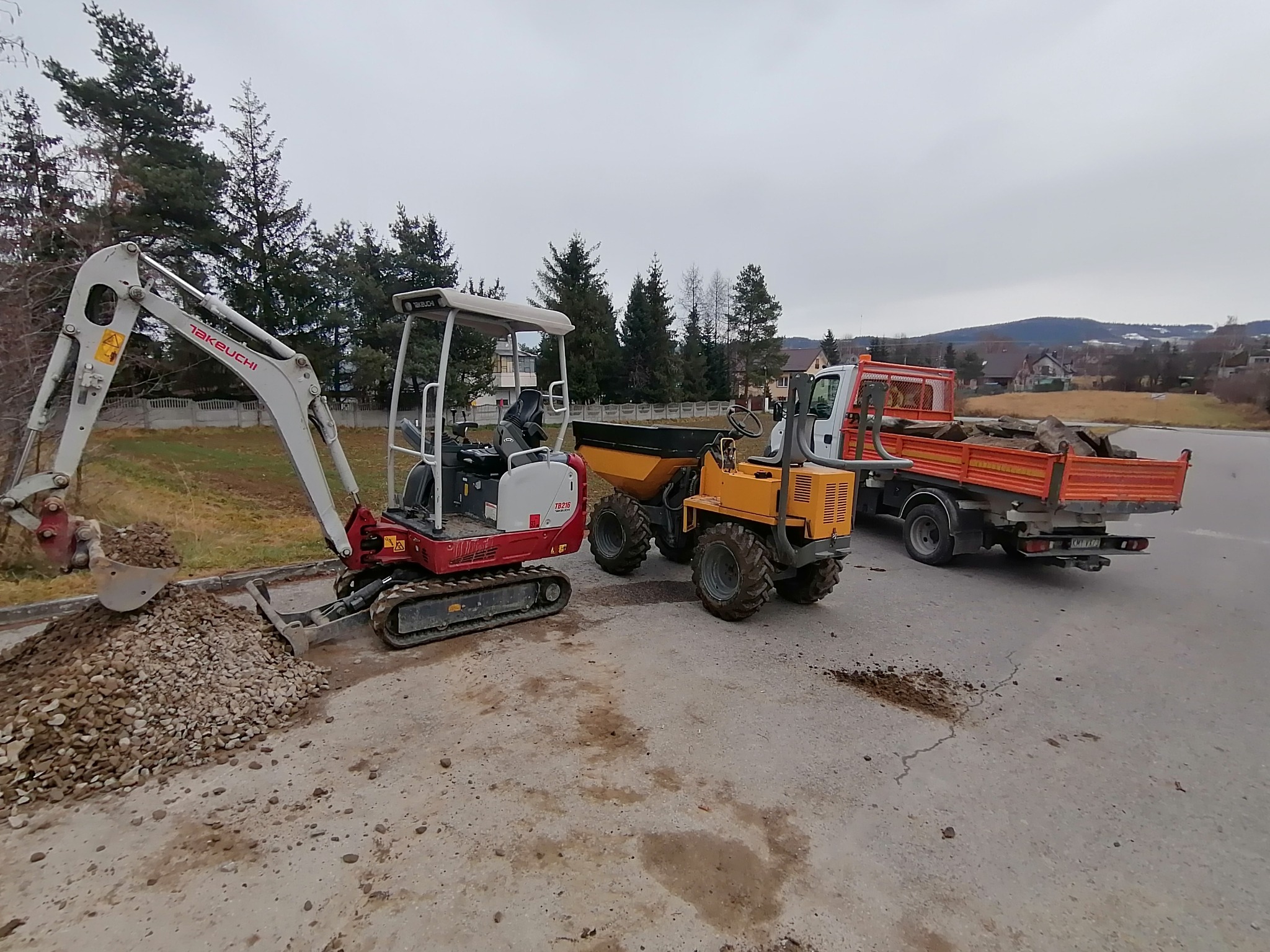 Mini koparka Takeuchi TB216 nabiera żwir, obok wywrotka i żółty wozidło budowlane, w tle zabudowa jednorodzinna i zalesione wzgórza.