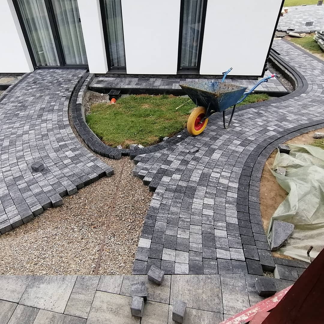 Trwa układanie szarej kostki brukowej w łukowaty wzór przed nowoczesnym domem z białymi ścianami i czarnymi oknami; widoczny fragment trawnika, taczka z żółtym kołem i rozsypany żwir.