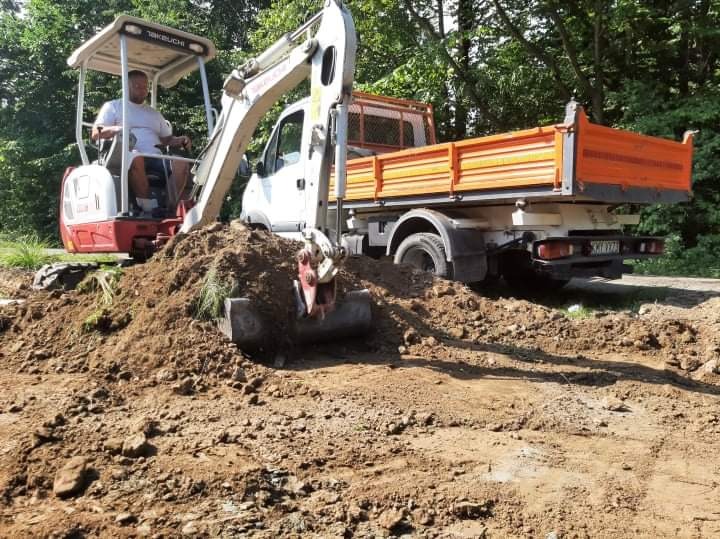Operator w minikoparce marki Takeuchi, obok wywrotka z pomarańczową skrzynią, prace ziemne w terenie zielonym.