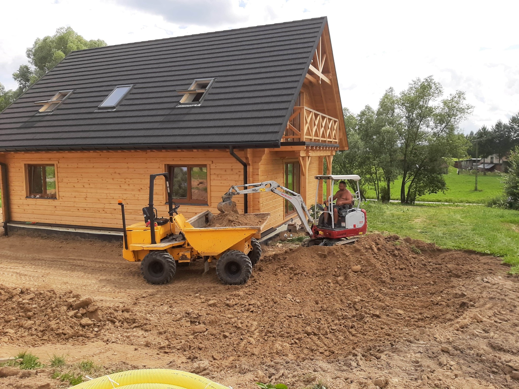 Żółta wywrotka wypełniona ziemią i mała koparka Takeuchi obok drewnianego domu z ciemnym dachem, przygotowanie terenu pod budowę, widoczne rury drenarskie.