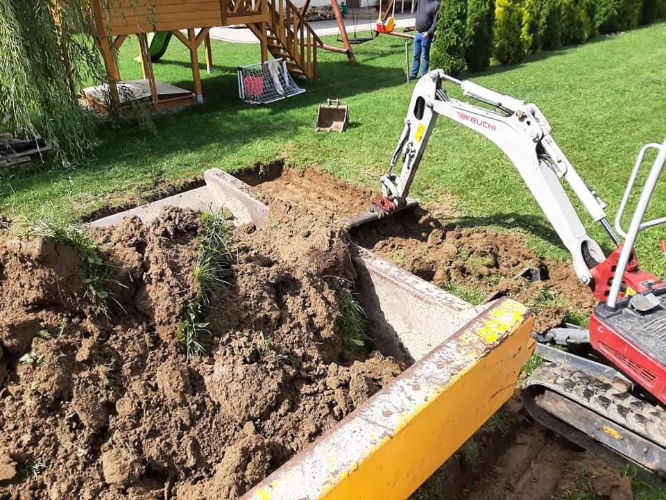 Mini koparka Takeuchi podczas prac ziemnych na trawniku, widoczny wykop z betonowym elementem, w tle plac zabaw i osoba.