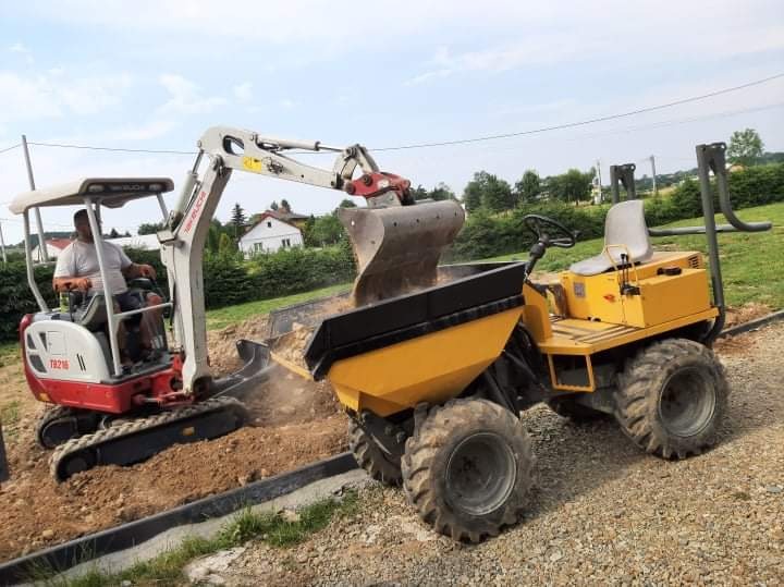 Mini koparka z operatorem wysypuje ziemię do wywrotki na terenie budowy z widocznymi krawężnikami