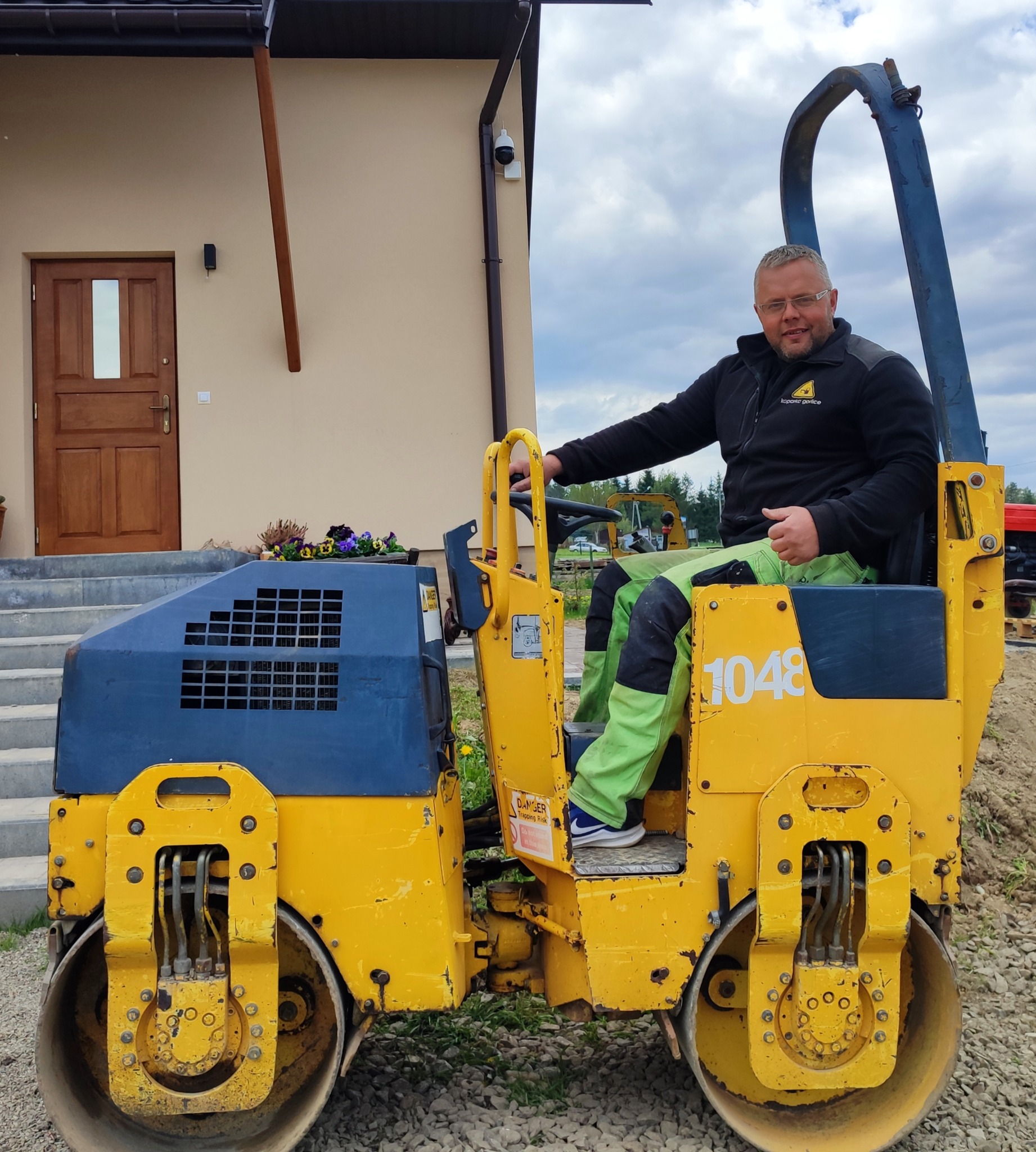 Operator siedzi na żółto-niebieskim walcu drogowym marki Dynapac obok nowo wybudowanego domu z brązowymi drzwiami wejściowymi i schodami.