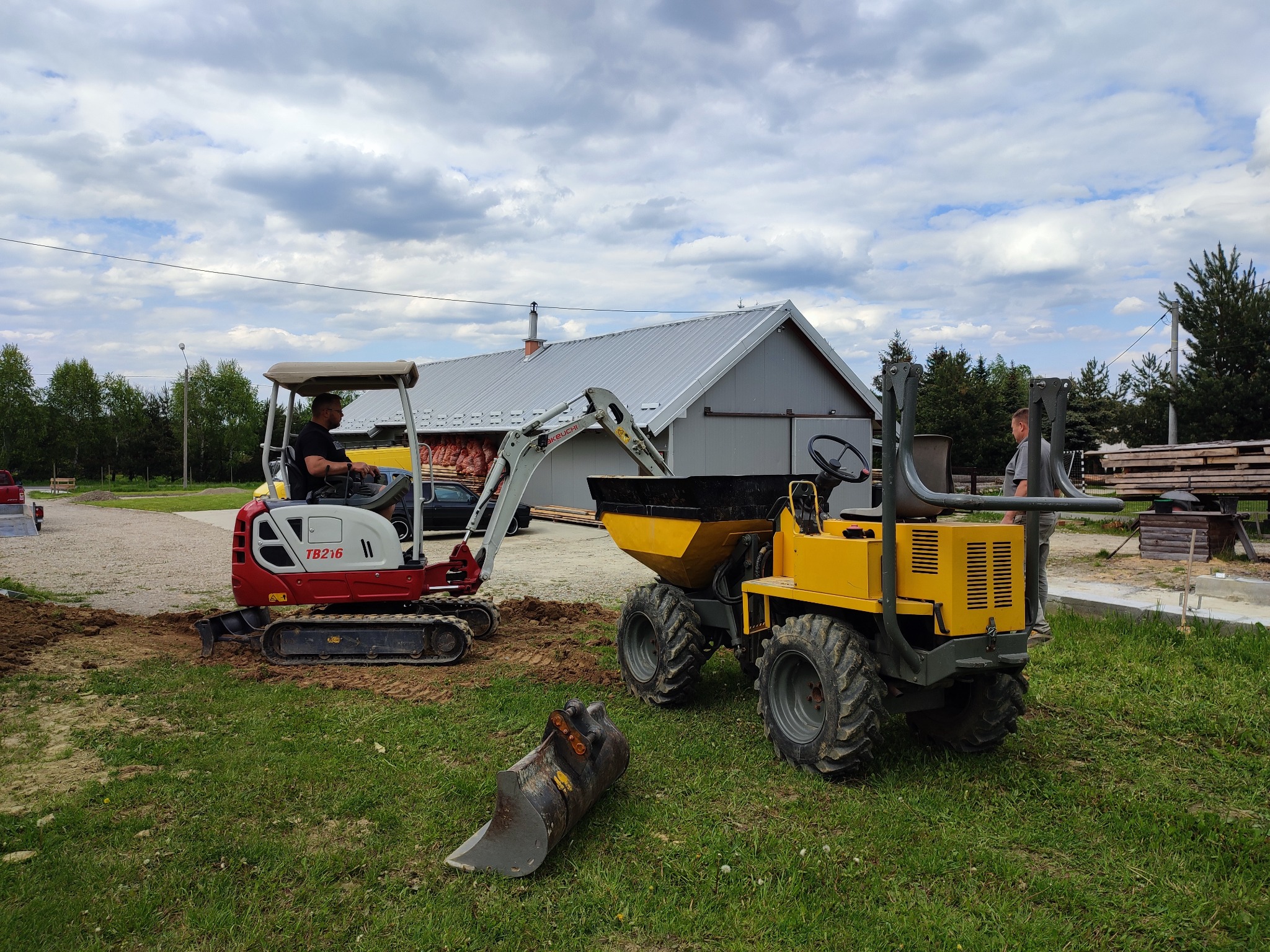 Czerwono-biała mini koparka Takeuchi TB216 z operatorem i żółta wywrotka na trawiastym terenie, w tle budynek z szarą blachą, fragment wykopu i dodatkowy osprzęt.