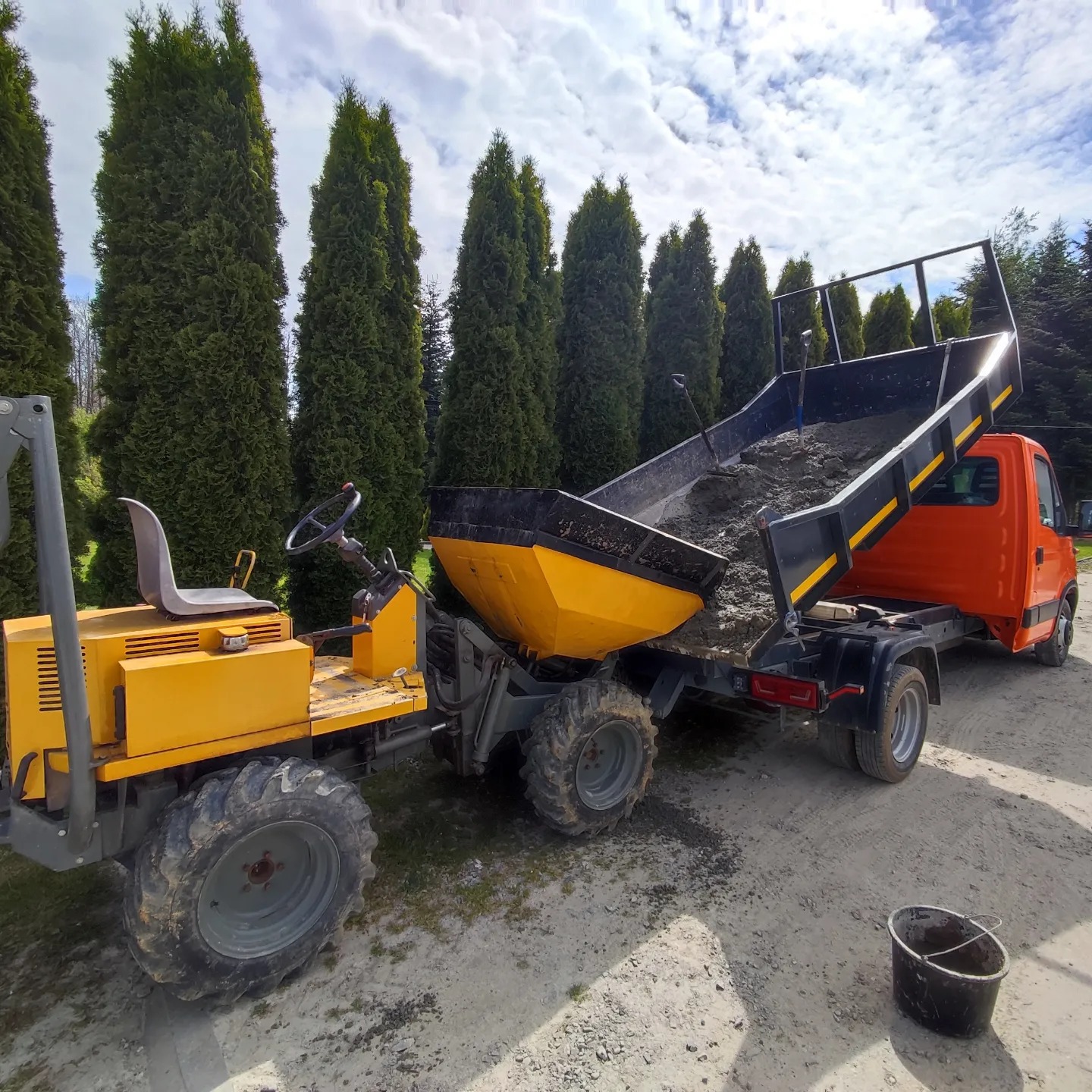 Pomarańczowa ciężarówka z żółtą przyczepą wyładowuje ziemię na placu budowy, w tle rząd wysokich tuj.