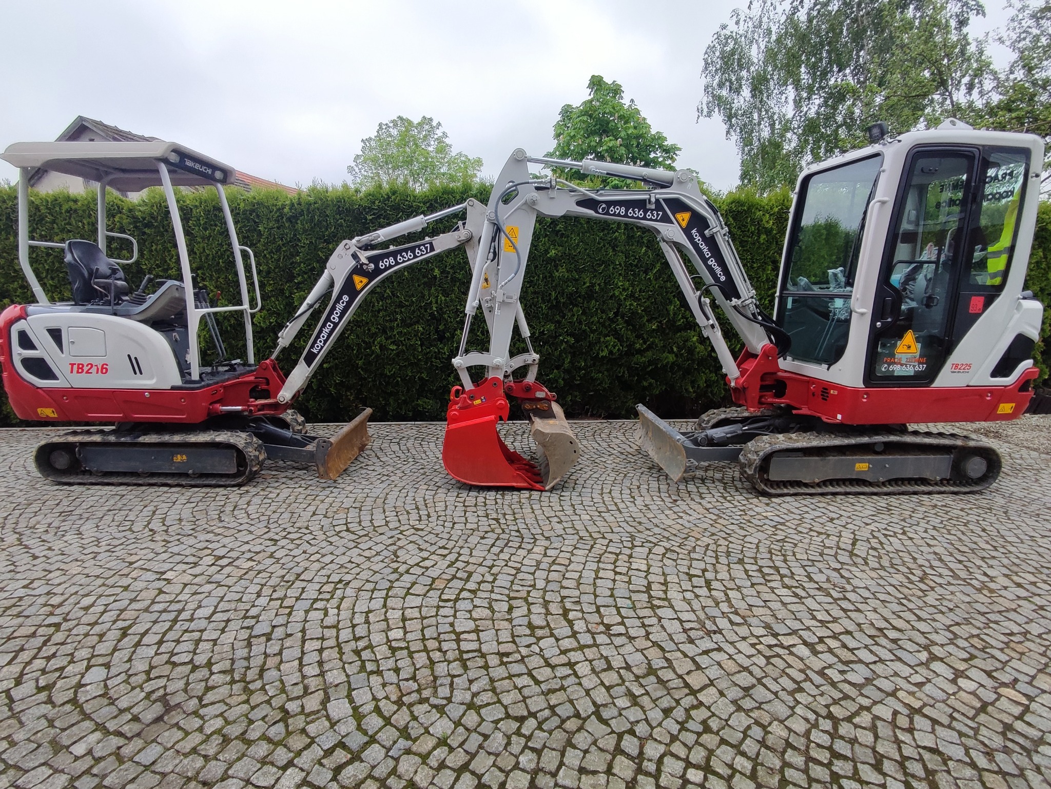 Dwie mini koparki Takeuchi, modele TB216 i TB225, zaparkowane na kostce brukowej przed żywopłotem, z widocznymi numerami telefonów kontaktowych na ramionach koparek.