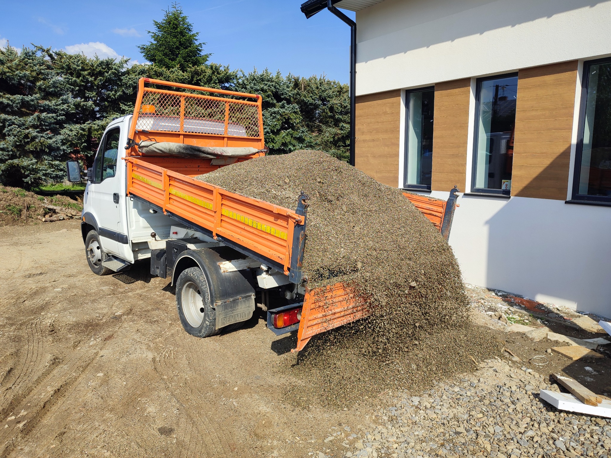 Pomarańczowo-biała wywrotka wysypuje żwir na plac budowy przy budynku z brązowymi panelami elewacyjnymi.