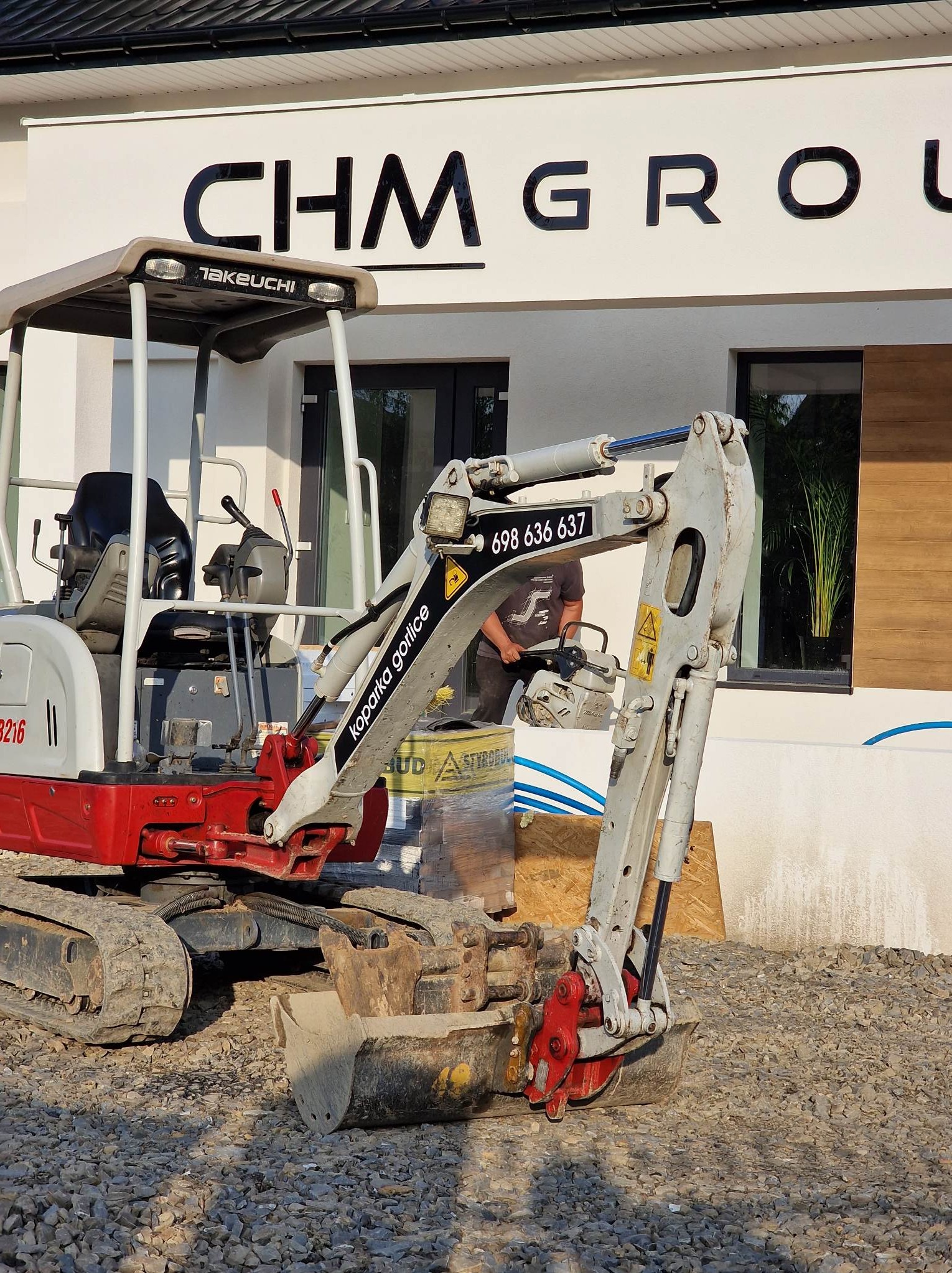 Kompaktowa, czerwono-szara koparka Takeuchi na żwirowym podłożu przed nowoczesnym budynkiem z napisem CHM GROUP, w tle widoczny pracownik z piłą.