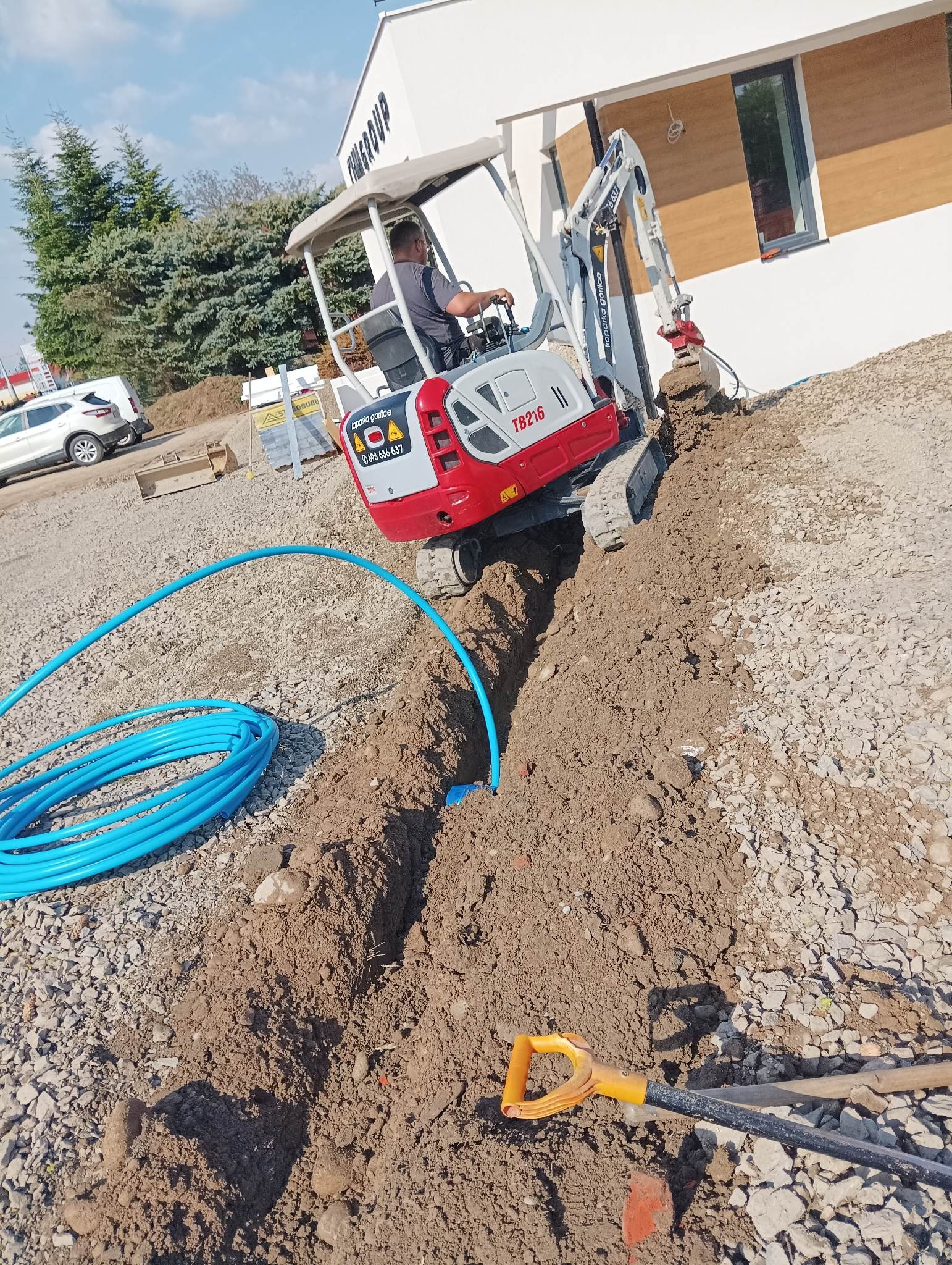 Mini koparka TB216 w trakcie wykonywania wykopu pod niebieskie rury na terenie budowy, widoczna łopata i budynek firmy w tle.