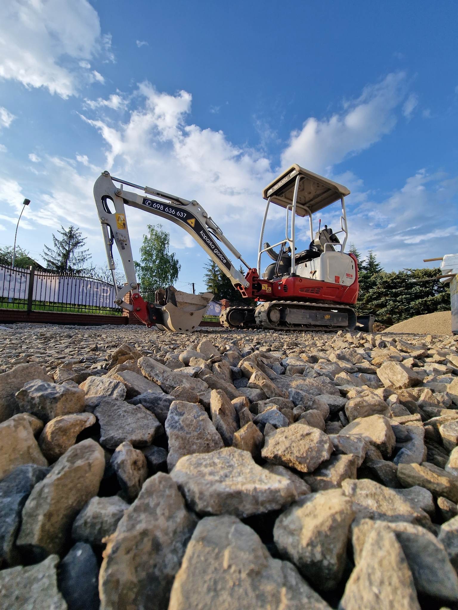 Czerwono-biała minikoparka Takeuchi TB216 stoi na warstwie tłucznia; w tle ogrodzenie z metalowych prętów i błękitne niebo z chmurami.