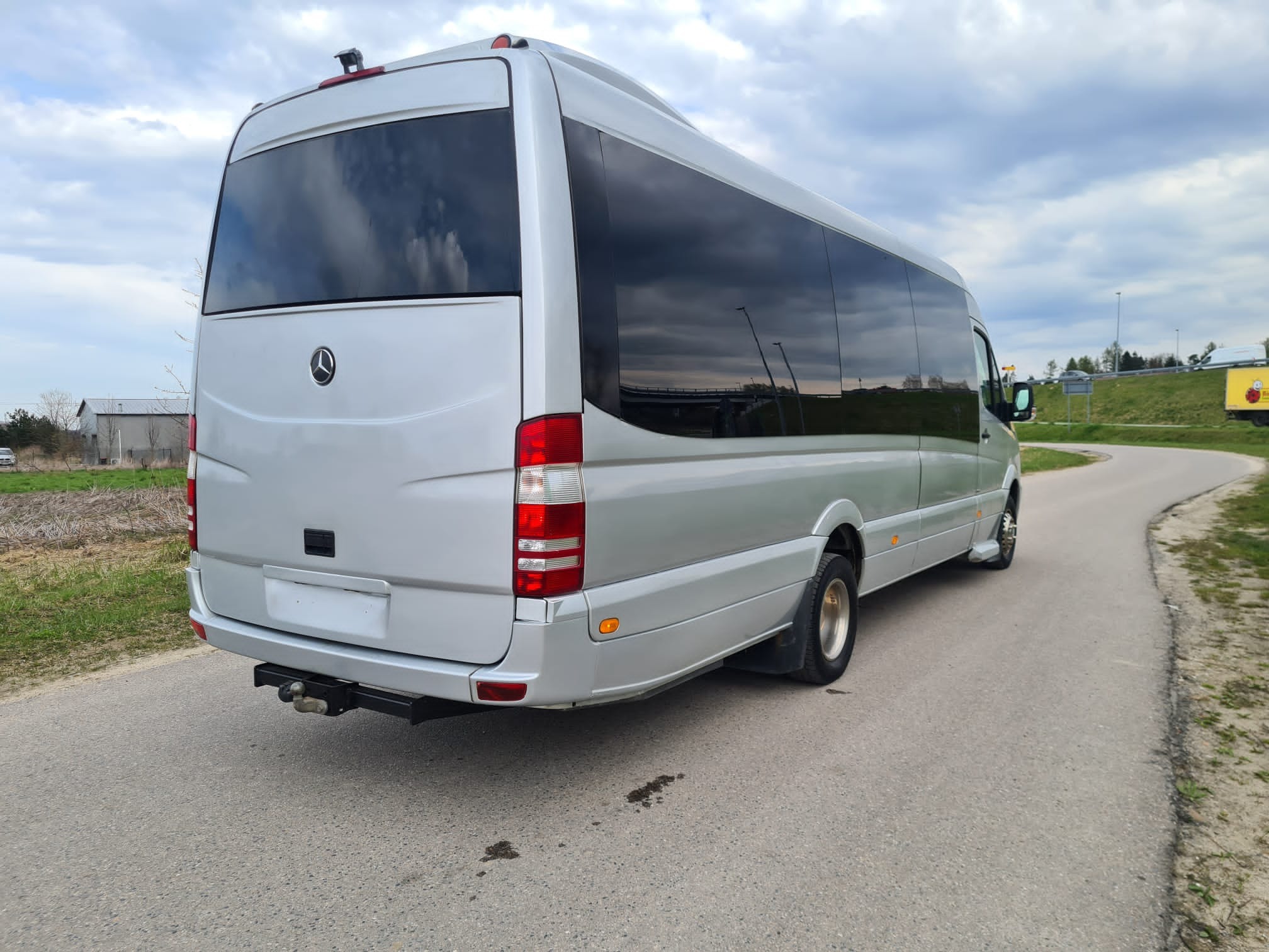 Srebrny bus Mercedes Sprinter z hakiem holowniczym, widok z tyłu na asfaltowej drodze w pochmurny dzień.