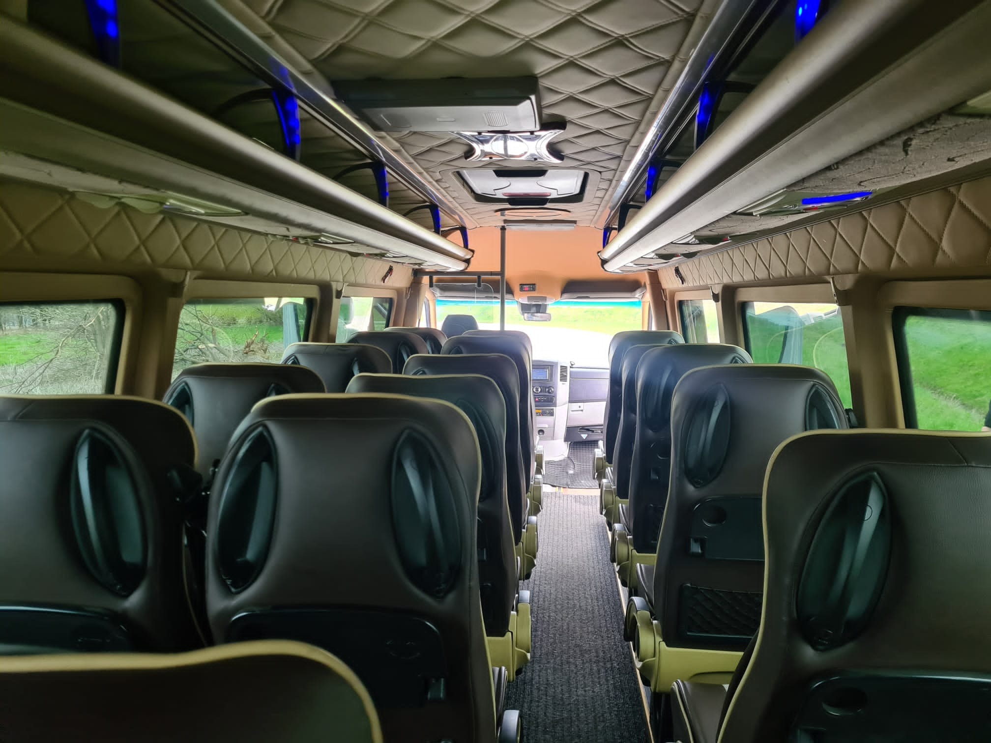 Wnętrze luksusowego busa z brązowymi skórzanymi siedzeniami, oświetleniem LED i pikowaną podsufitką. Widok na kabinę kierowcy przez przejście między rzędami siedzeń.