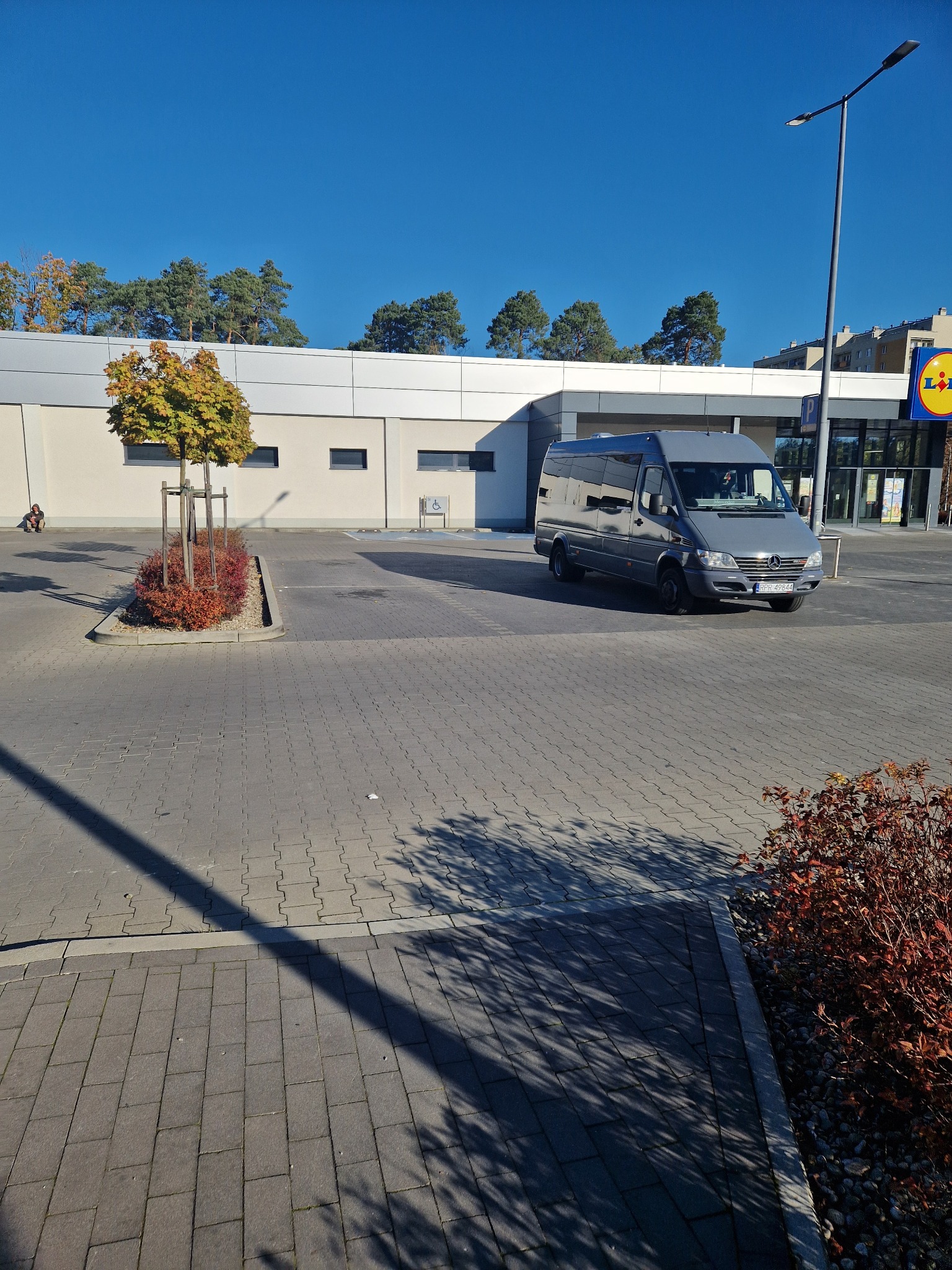 Szary bus Mercedes-Benz Sprinter zaparkowany na parkingu przed sklepem Lidl, widoczny numer rejestracyjny RPR 49644, w tle błękitne niebo i drzewa.