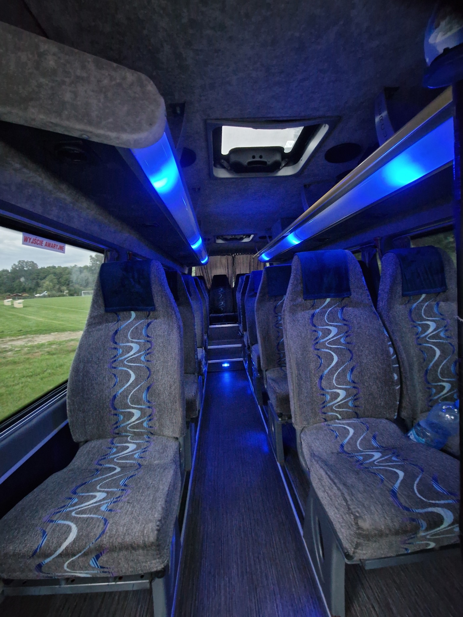 Wnętrze autobusu z szarymi fotelami z niebieskim wzorem, podświetlonymi niebieskim światłem LED, widok z perspektywy pasażera w kierunku przodu pojazdu.
