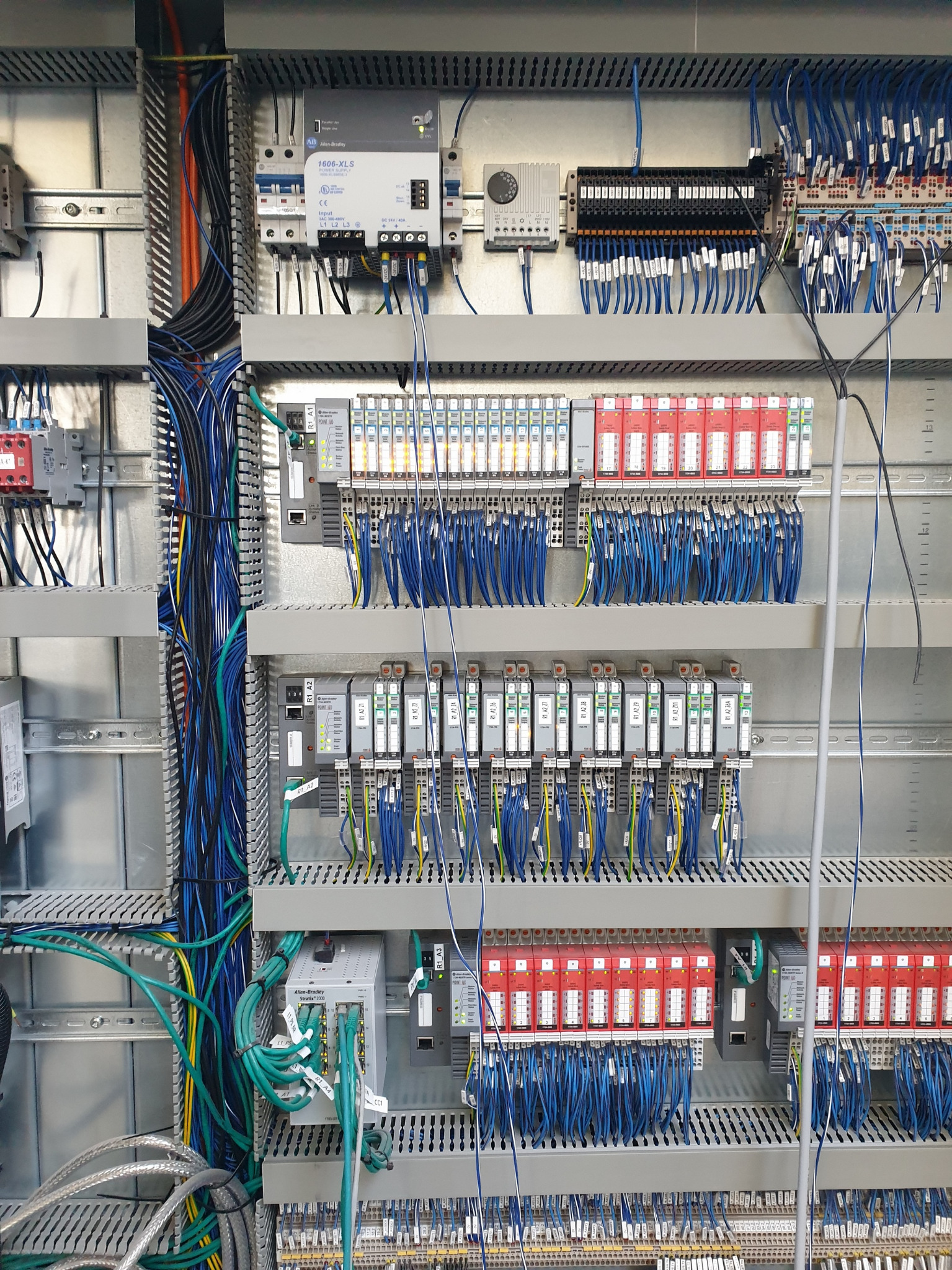 Panel sterowania elektrycznego z modułami Allen-Bradley, widoczne liczne kable w kolorach niebieskim, zielonym i czarnym, oraz moduły z oznaczeniami R1 A2, R1 A3, R1 A4, Stratix 2000 i 1606-XLS.