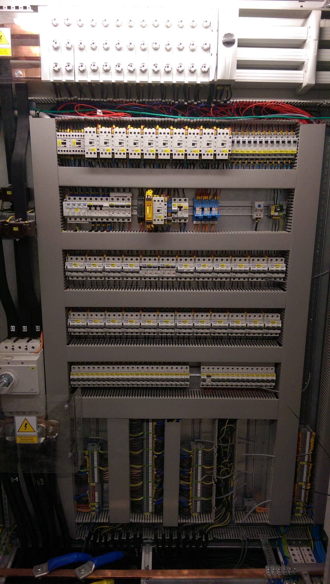 Rozdzielnica elektryczna z licznymi zabezpieczeniami i okablowaniem, widoczne bezpieczniki, przekaźniki i listwy zaciskowe, z niebieskimi szczypcami u dołu.