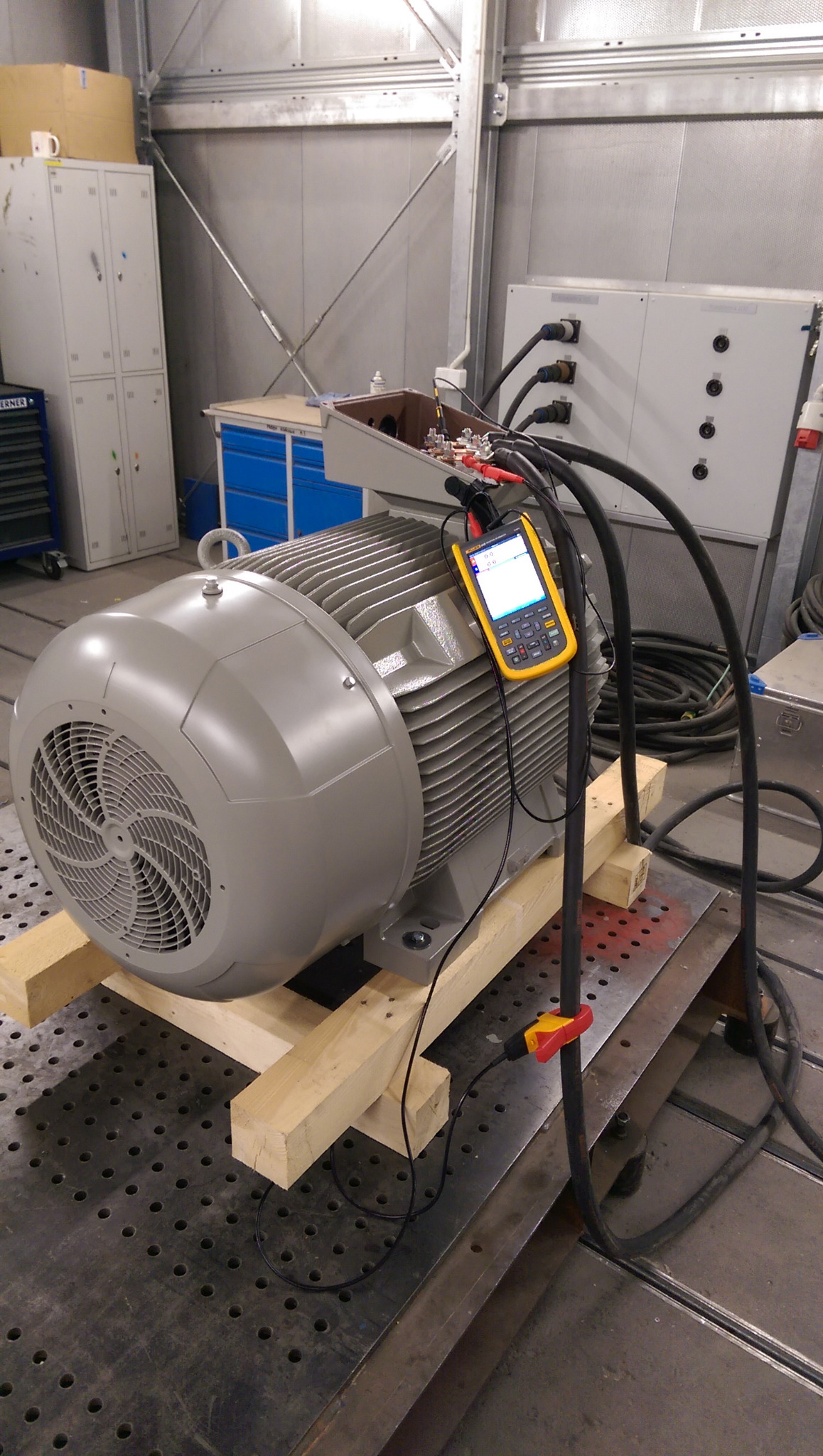 Duży silnik elektryczny na drewnianych podporach, podłączony do przewodów i miernika z wyświetlaczem, w tle szafki i skrzynka przyłączeniowa.