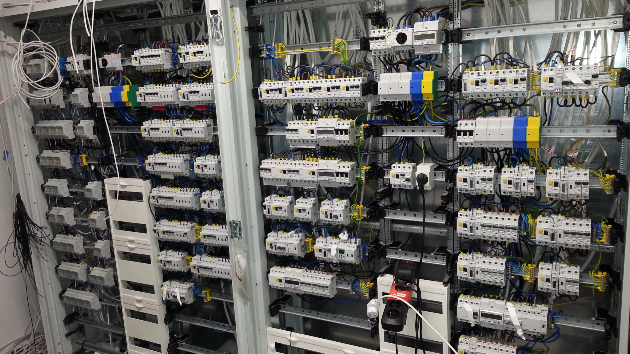 Rozdzielnia elektryczna z licznymi zabezpieczeniami, bezpiecznikami i przewodami, widoczne szyny montażowe i elementy oznaczone kolorami niebieskim i żółtym.