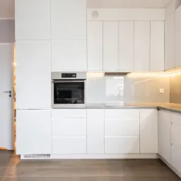 Minimalistyczna biała kuchnia z połyskującymi frontami, oświetleniem LED pod szafkami i otwartymi drzwiami do spiżarni.