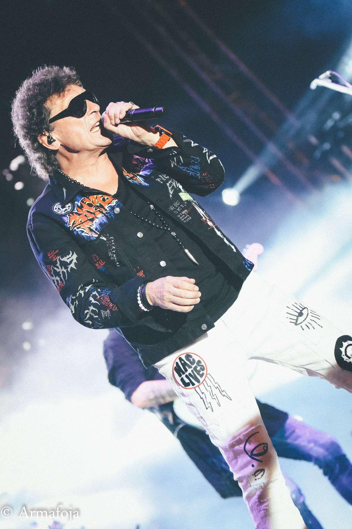 Piosenkarz z kręconymi włosami i w okularach przeciwsłonecznych, w czarnej kurtce z kolorowymi grafikami i białych spodniach z nadrukami, śpiewa do mikrofonu na scenie oświetlonej reflektorami.