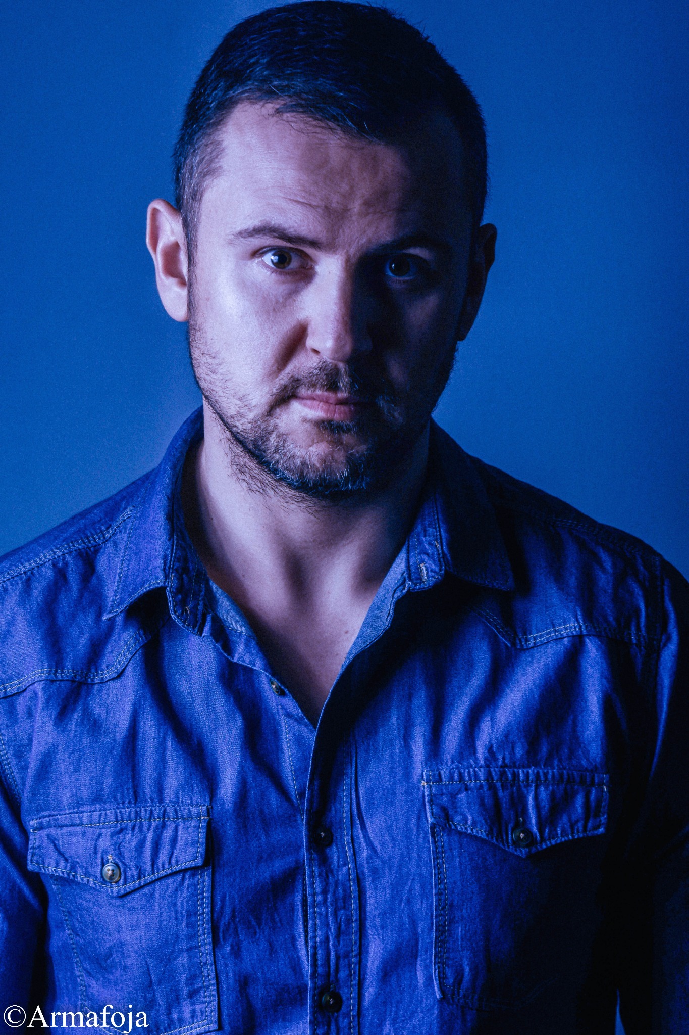 Portret mężczyzny w dżinsowej koszuli na niebieskim tle, oświetlonego chłodnym światłem.
