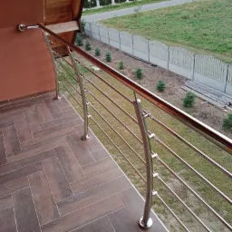 Balkon z balustradą nierdzewną o poziomych prętach, widok z góry na brązowe płytki podłogowe, w tle trawnik i betonowe ogrodzenie.