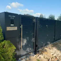 Nowoczesna, przesuwna brama wjazdowa w kolorze grafitowym z panelem kontrolnym, skrzynką na listy i długim, pionowym uchwytem ze stali nierdzewnej, zamontowana przy posesji z zielonymi krzewami...