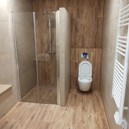 wykonczeniowka plytki regips szpachlowanie i inne - Nowoczesna łazienka z kabiną prysznicową ze szklanymi drzwiami, toaletą wiszącą z chromowanym przyciskiem spłukiwania oraz grzejnikiem drabinkowym, wykończona płytkami imitującymi drewno...
