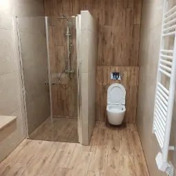 Nowoczesna łazienka z kabiną prysznicową ze szklanymi drzwiami, toaletą wiszącą z chromowanym przyciskiem spłukiwania oraz grzejnikiem drabinkowym, wykończona płytkami imitującymi drewno...