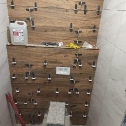 wykonczeniowka plytki regips szpachlowanie i inne - Ściana w trakcie układania płytek imitujących drewno, z krzyżykami dystansowymi, widoczna poziomica, drabina, preparat gruntujący i akcesoria glazurnicze.