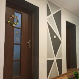 Wnętrze przedpokoju z brązowymi drzwiami wejściowymi ozdobionymi wiosennym wiankiem i geometrycznym malowaniem ściany w odcieniach szarości i bieli.