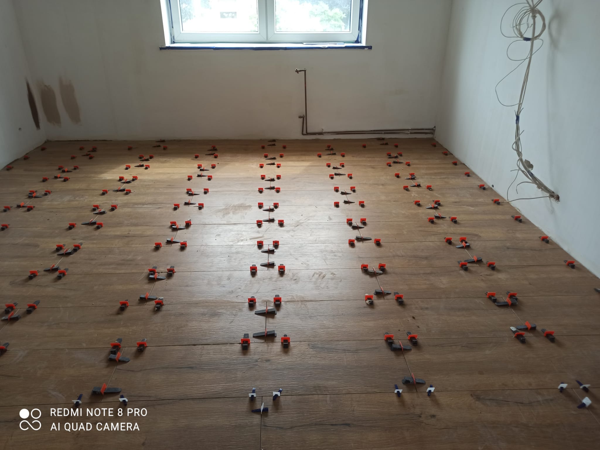 Układanie płytek podłogowych imitujących drewno z użyciem systemu poziomowania, widoczne krzyżyki dystansowe i kliny, surowy stan ścian i okno w tle.