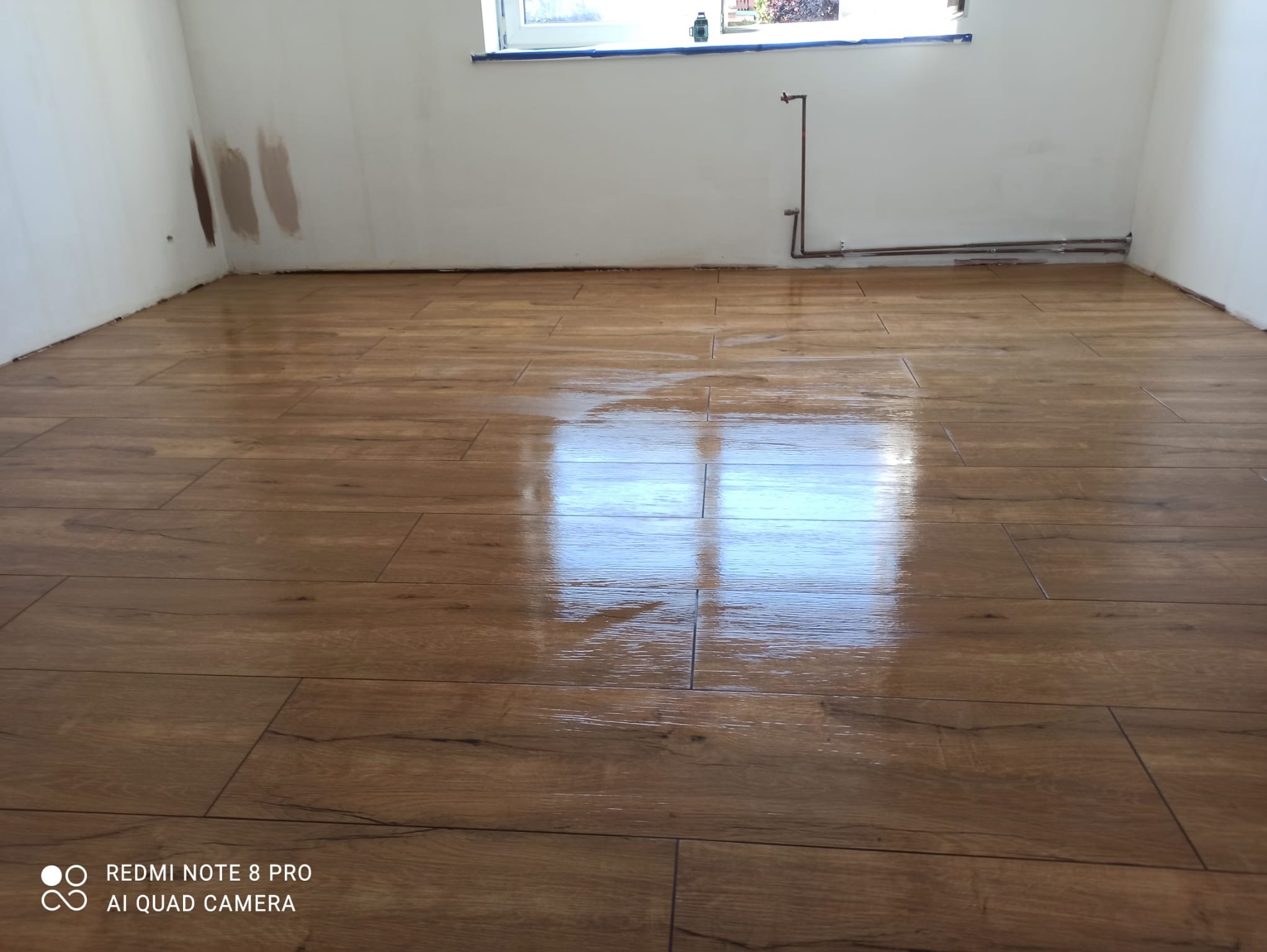 Świeżo położona, błyszcząca podłoga z paneli imitujących drewno w pustym pokoju z białymi ścianami. Na ścianie widoczne ślady po próbkach farb.