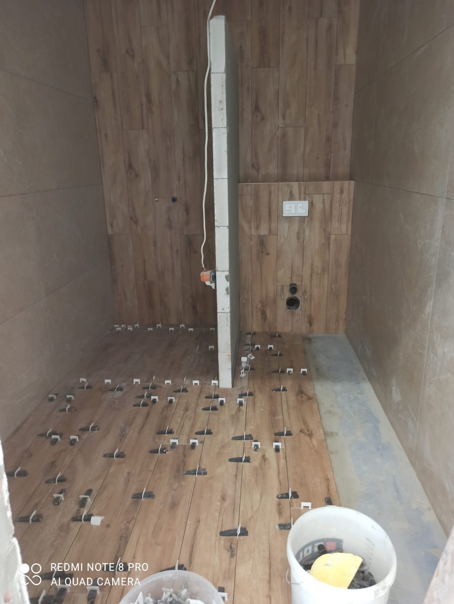 Łazienka w trakcie remontu: układanie drewnopodobnych płytek na podłodze z użyciem klipsów poziomujących, ściany wyłożone płytkami, widoczny fragment ściany działowej z instalacją elektryczną.