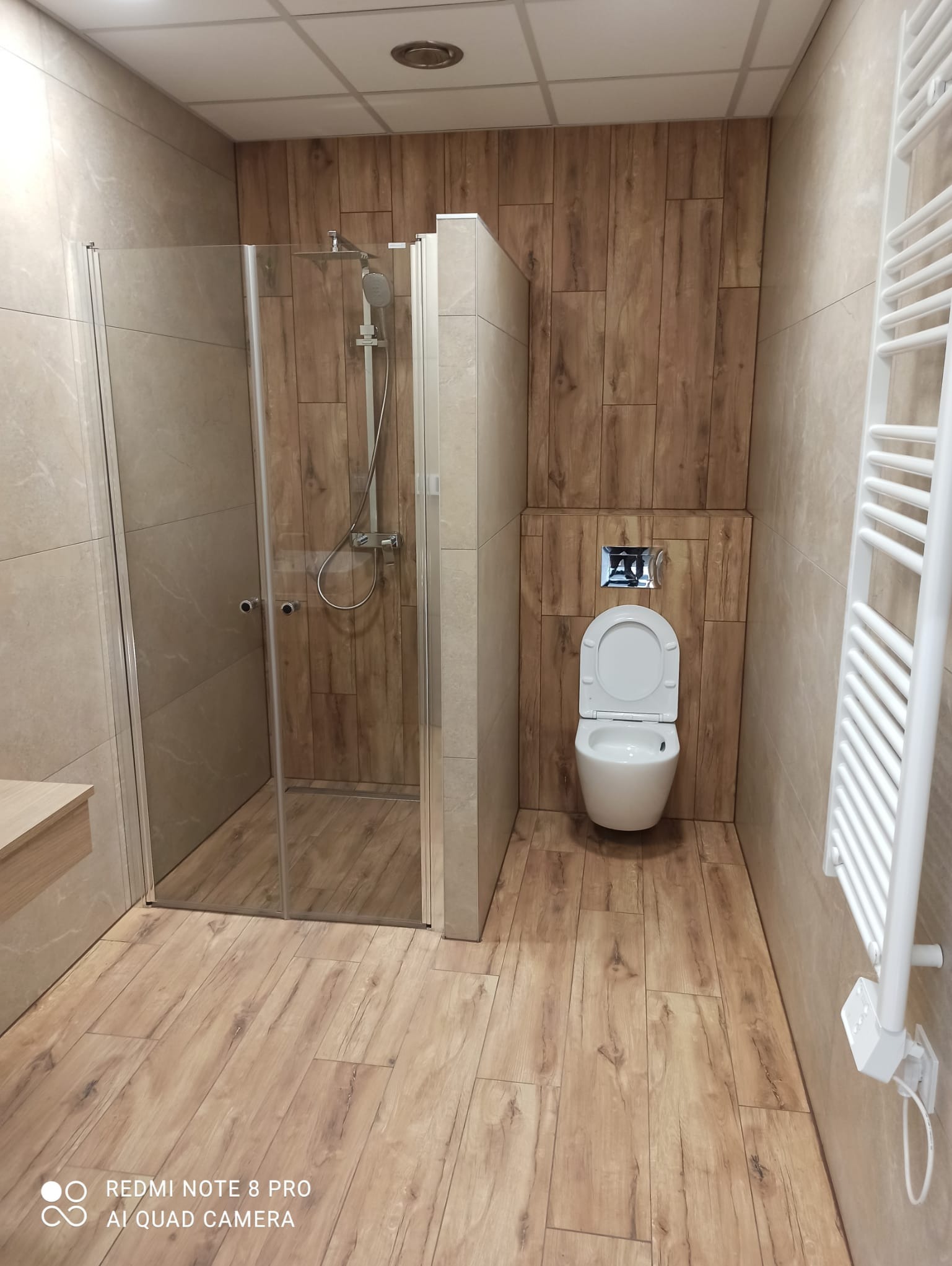 Nowoczesna łazienka z kabiną prysznicową ze szklanymi drzwiami, toaletą wiszącą z chromowanym przyciskiem spłukiwania oraz grzejnikiem drabinkowym, wykończona płytkami imitującymi drewno...