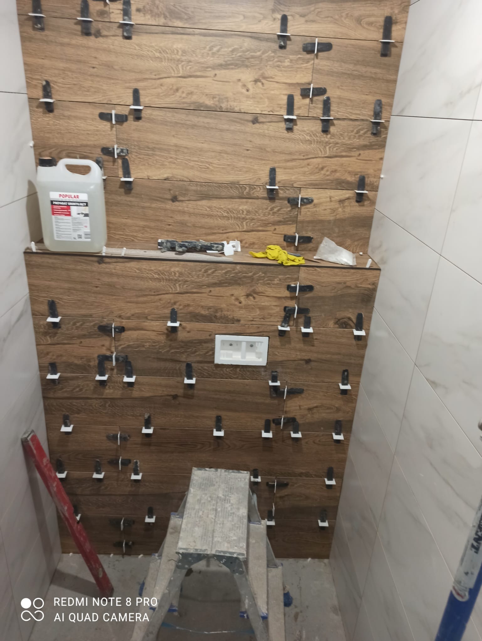 Ściana w trakcie układania płytek imitujących drewno, z krzyżykami dystansowymi, widoczna poziomica, drabina, preparat gruntujący i akcesoria glazurnicze.