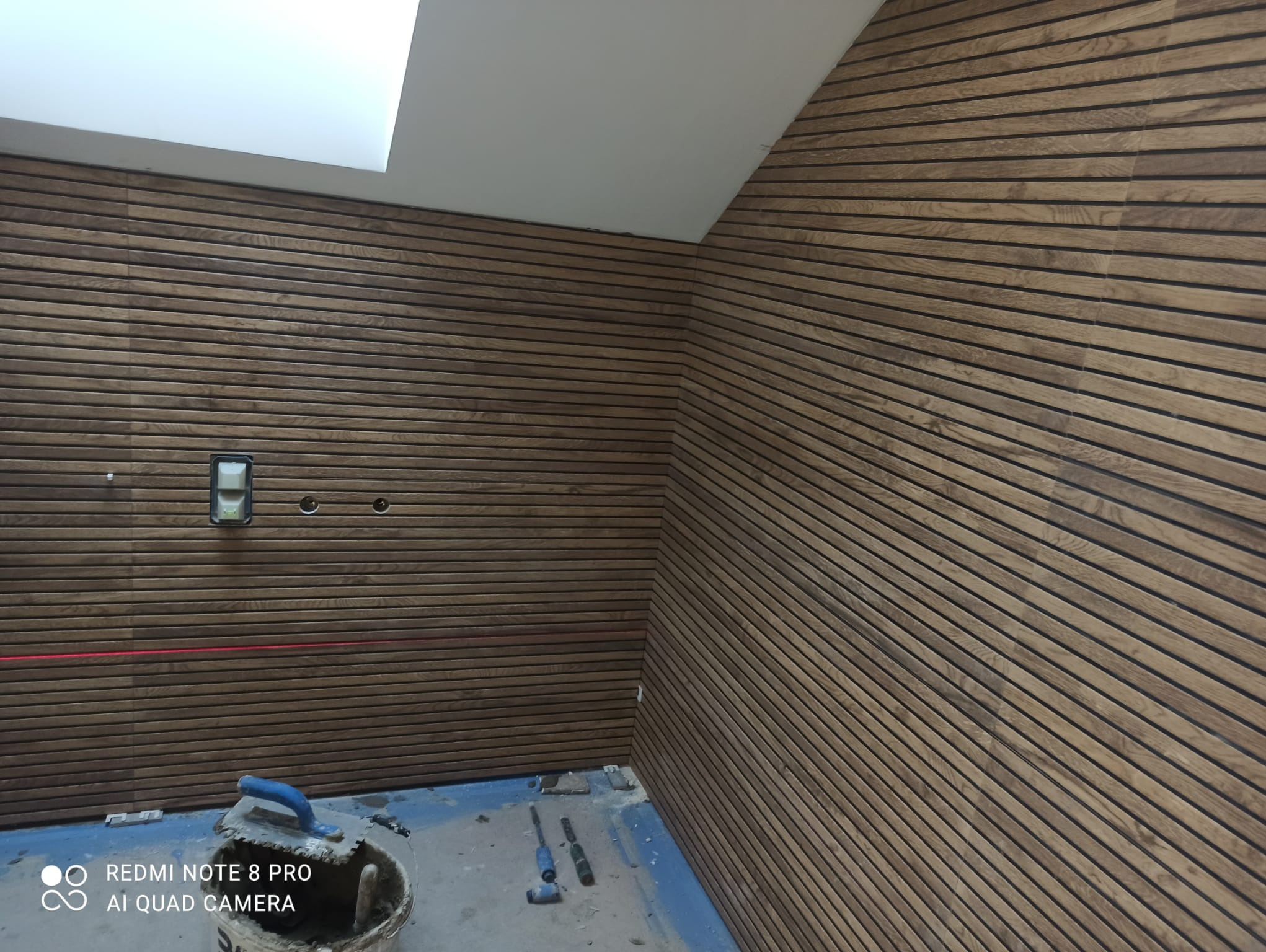 Drewniana boazeria lamelowa na ścianie w trakcie montażu, widoczny laser krzyżowy i narzędzia glazurnicze na podłodze, sufit z oknem dachowym.