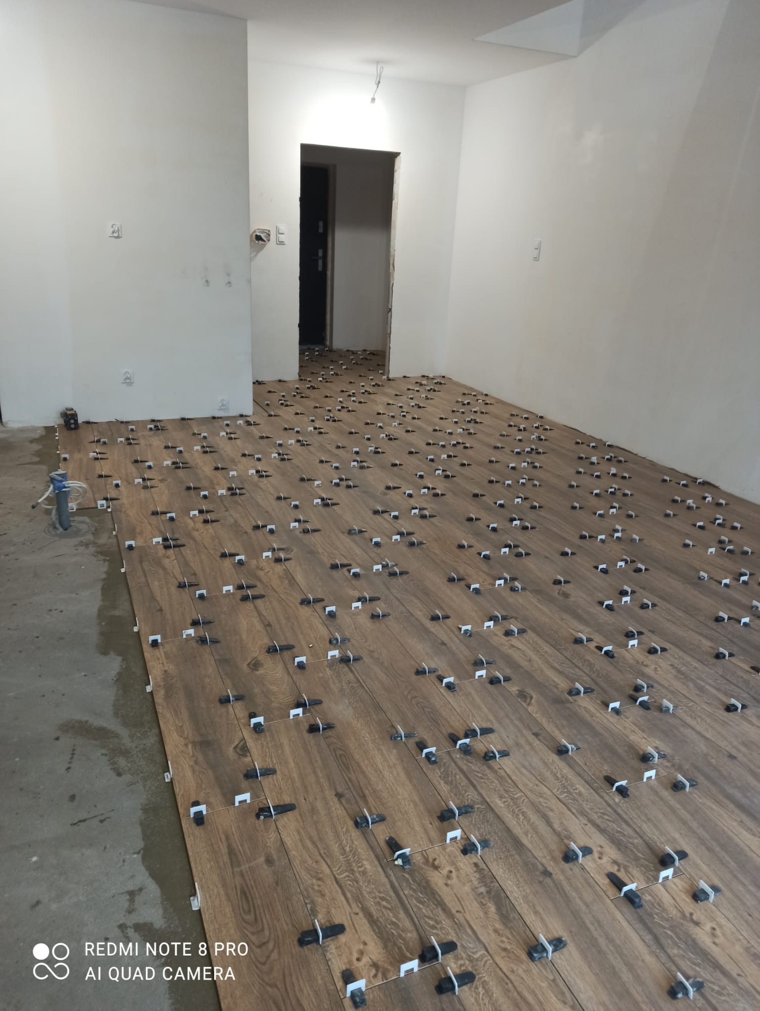 Układanie płytek imitujących drewno na podłodze w nowym mieszkaniu z użyciem klipsów poziomujących, widoczny stan surowy ścian i podłogi.