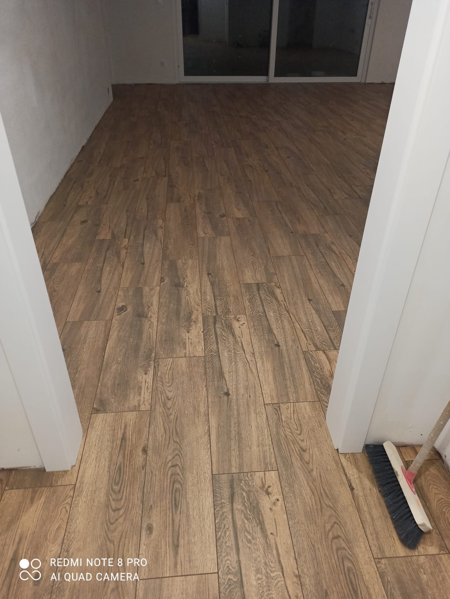 Ułożona podłoga z paneli imitujących drewno, widok z perspektywy wejścia do pomieszczenia, przy futrynie stoi zmiotka.