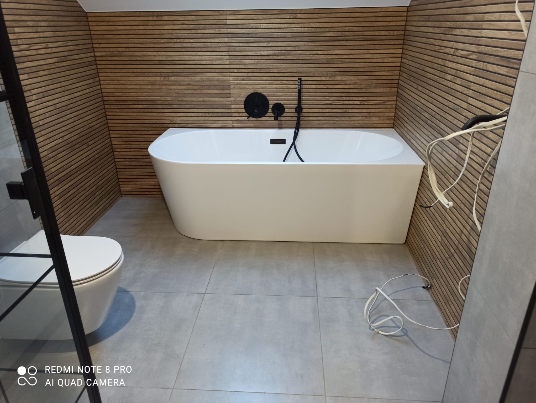 Nowoczesna łazienka z wolnostojącą wanną, drewnianą okładziną ścienną i czarną armaturą, widoczne niedokończone prace instalacyjne.