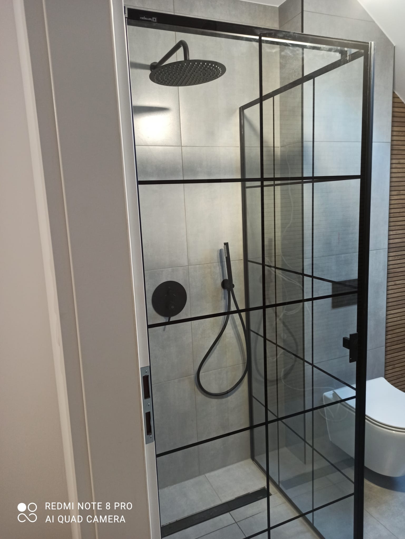 Nowoczesna kabina prysznicowa z czarnymi profilami i deszczownicą w łazience wykończonej szarymi płytkami i drewnianymi panelami.