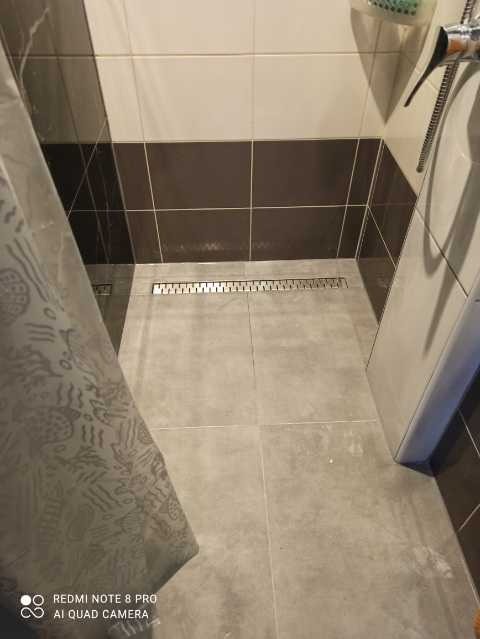 Szara, wykafelkowana podłoga prysznica z odpływem liniowym, widok częściowo zasłonięty przez wzorzystą zasłonę prysznicową, z fragmentem baterii i płytek ściennych w odcieniach brązu i beżu.