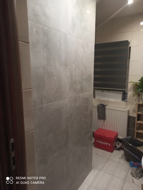 Wykończona ściana łazienki z szarych płytek imitujących beton, widoczny fragment grzejnika, rolety w oknie i czerwona skrzynka narzędziowa Hilti w tle.