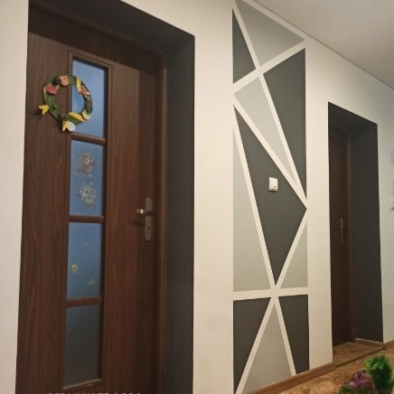 Wnętrze przedpokoju z brązowymi drzwiami wejściowymi ozdobionymi wiosennym wiankiem i geometrycznym malowaniem ściany w odcieniach szarości i bieli.