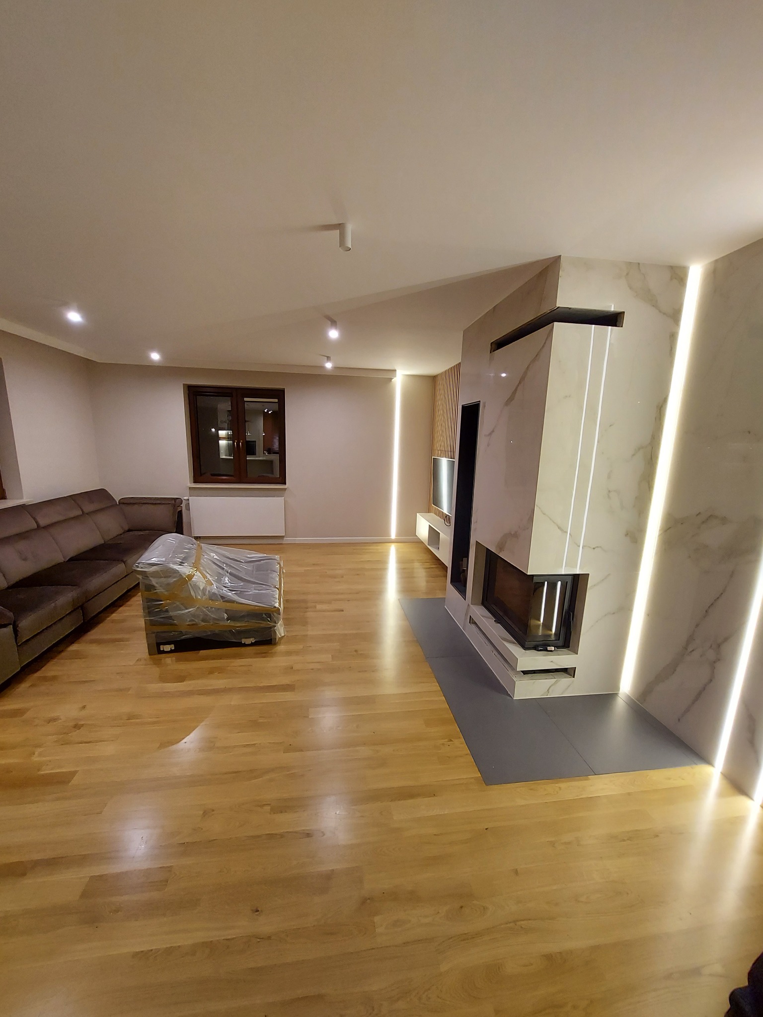 Nowoczesny salon z drewnianą podłogą, marmurowym kominkiem z podświetleniem LED i częściowo zakrytym meblem folią ochronną.