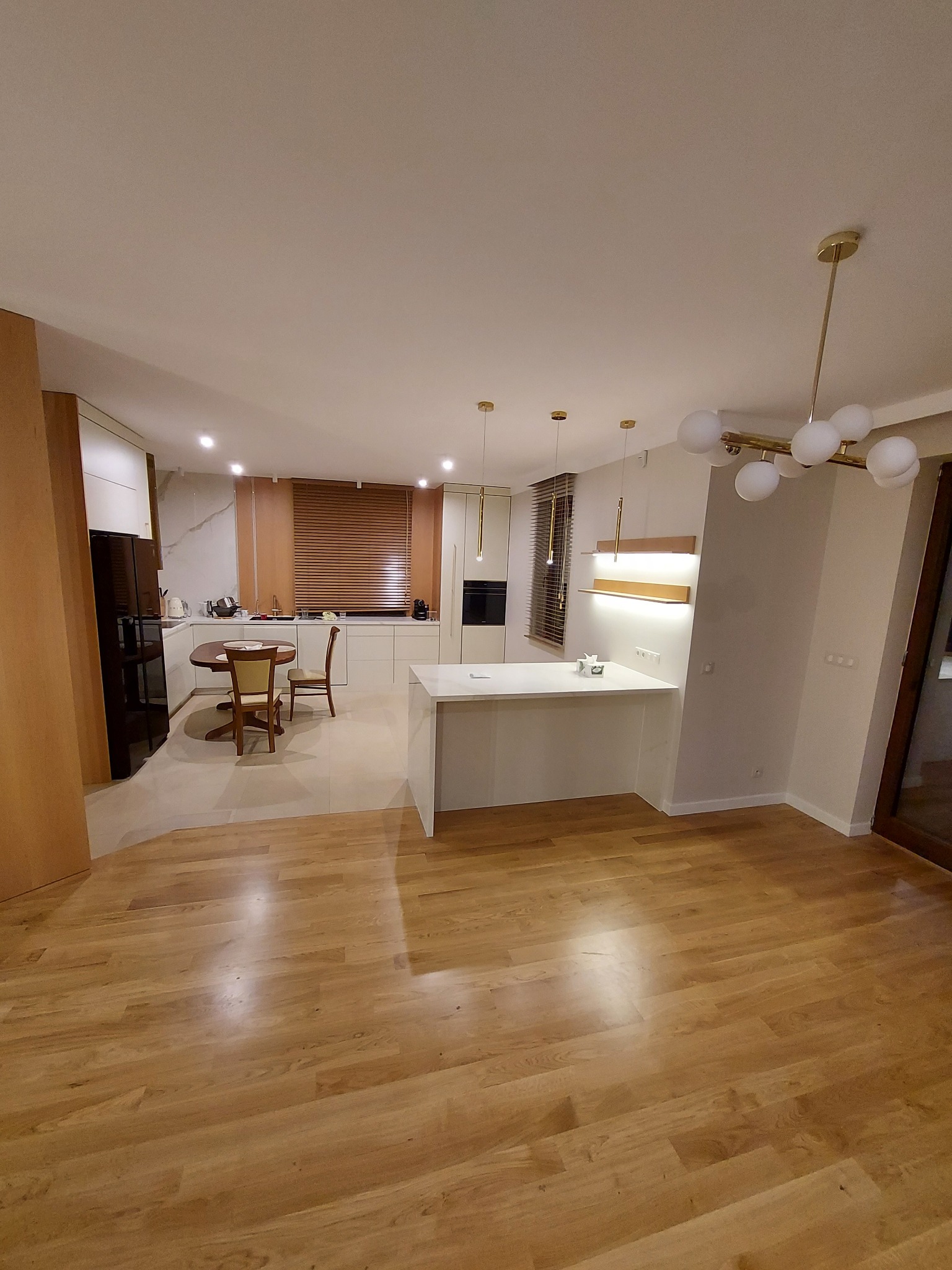 Nowoczesna, jasna kuchnia z drewnianą podłogą i wyspą kuchenną, oświetlona lampami wiszącymi i żaluzjami.