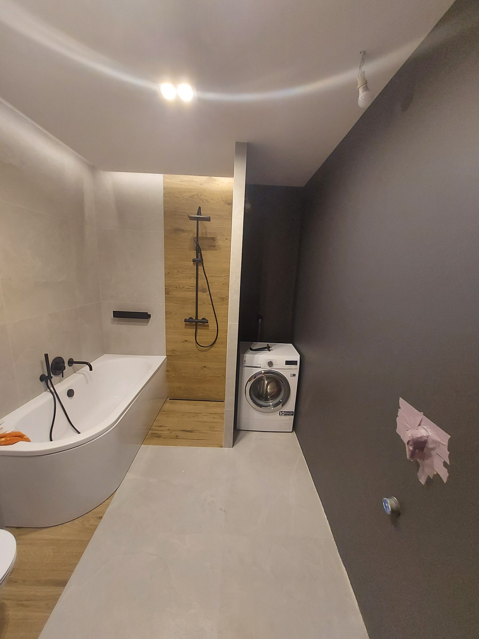 Nowoczesna łazienka z wanną, drewnopodobną ścianką prysznicową z czarną armaturą, pralką i szarą ścianą z widocznymi śladami po instalacji.